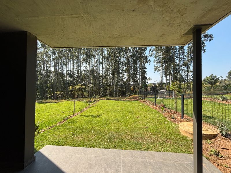 A covered patio area featuring tiled flooring and support columns, overlooking a private garden with a wire mesh fence and mature trees.
