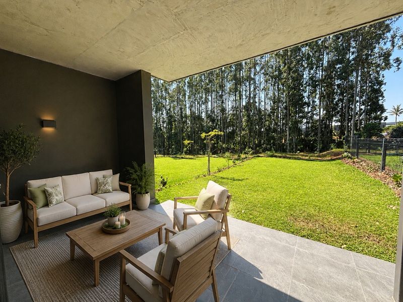 A covered patio featuring modern tiled flooring and recessed wall lighting, overlooking a lush green garden and forest backdrop.