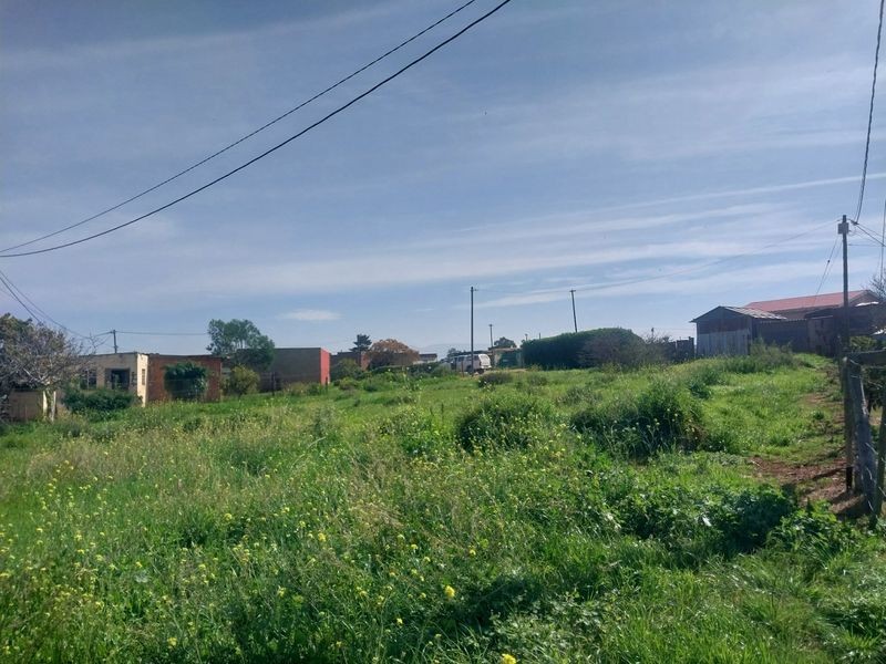 A view of a vacant residential plot featuring overgrown grass, utility poles, and overhead power lines with surrounding neighborhood structures visible in the distance.