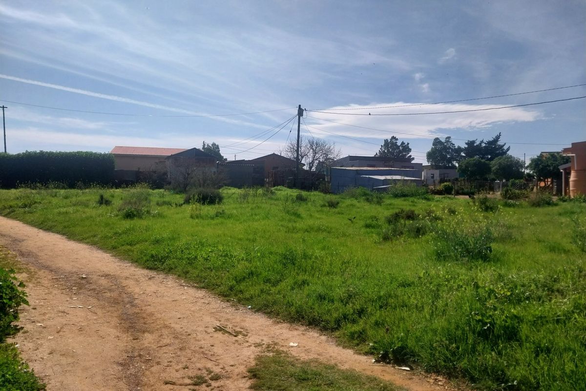 A vacant residential plot featuring a dirt access road, grassy terrain, and overhead utility infrastructure.