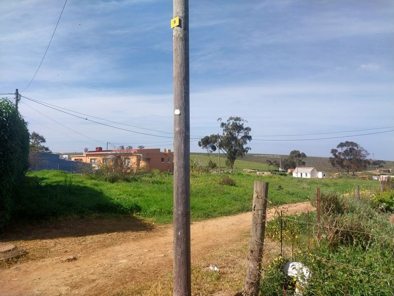 A view of a vacant residential plot featuring a utility pole, wire fencing, and a dirt access path with surrounding landscape.