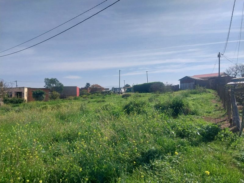 A vacant residential plot featuring overgrown grass, a wooden boundary fence, and overhead utility lines, situated in a residential neighborhood.