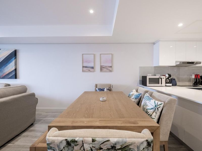 A modern open-plan living and dining area featuring sleek white kitchen cabinetry, stone countertops, recessed ceiling lighting, and neutral tiled flooring.