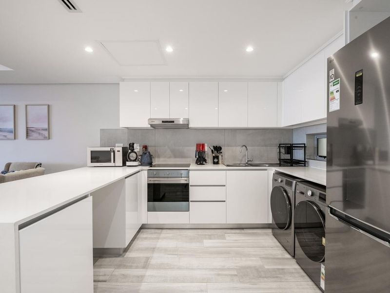 Modern kitchen featuring sleek white cabinetry, stone countertops, integrated appliances, and a clean tiled backsplash in a bright, open-plan layout.
