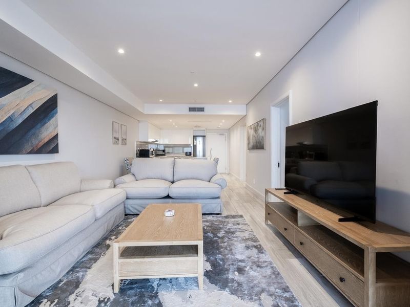 Modern open-plan living area featuring light timber-look flooring, recessed downlighting, and integrated ducted air conditioning, flowing seamlessly into a contemporary kitchen space.