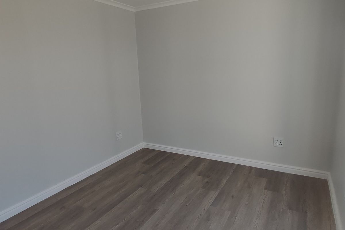 A bright, empty bedroom featuring modern laminate flooring, neutral walls, and a large window providing natural light.