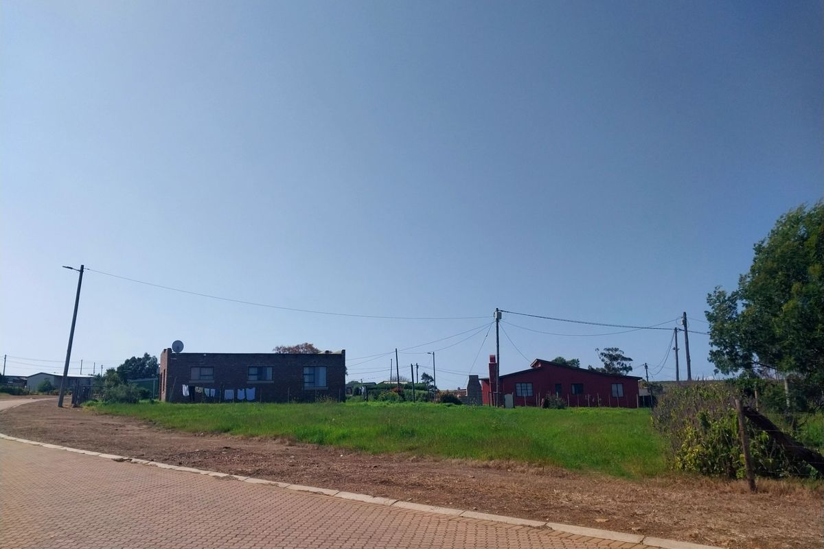 A street-level view of a residential area featuring a paved road, utility infrastructure, and open grass verge leading towards existing properties.