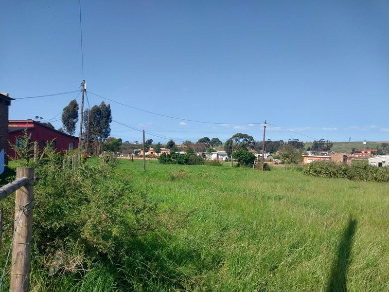 A view of a vacant residential plot featuring grassy terrain, perimeter wire fencing, and overhead utility infrastructure with surrounding neighborhood buildings in the distance.