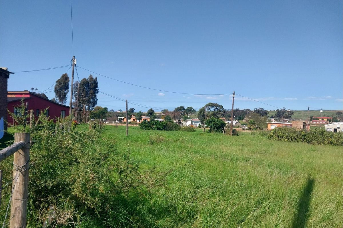 A view of a vacant residential plot featuring grassy terrain, perimeter wire fencing, and overhead utility infrastructure with surrounding neighborhood buildings in the distance.