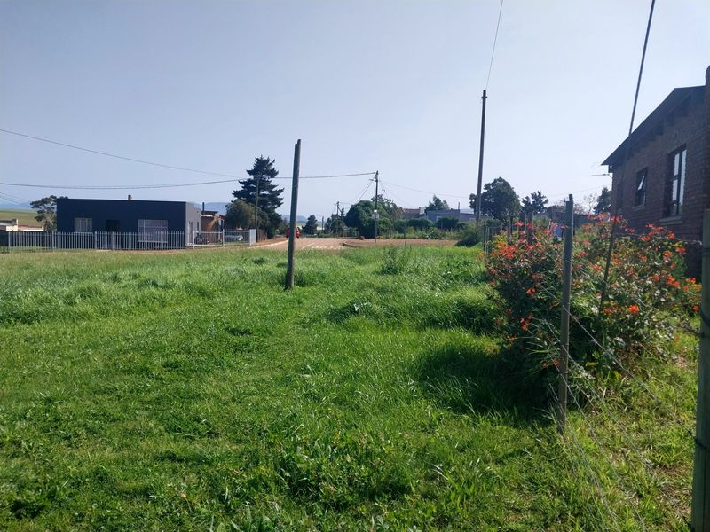 A view of a vacant residential plot featuring grassy terrain, perimeter wire fencing, and utility infrastructure with neighboring properties visible in the background.