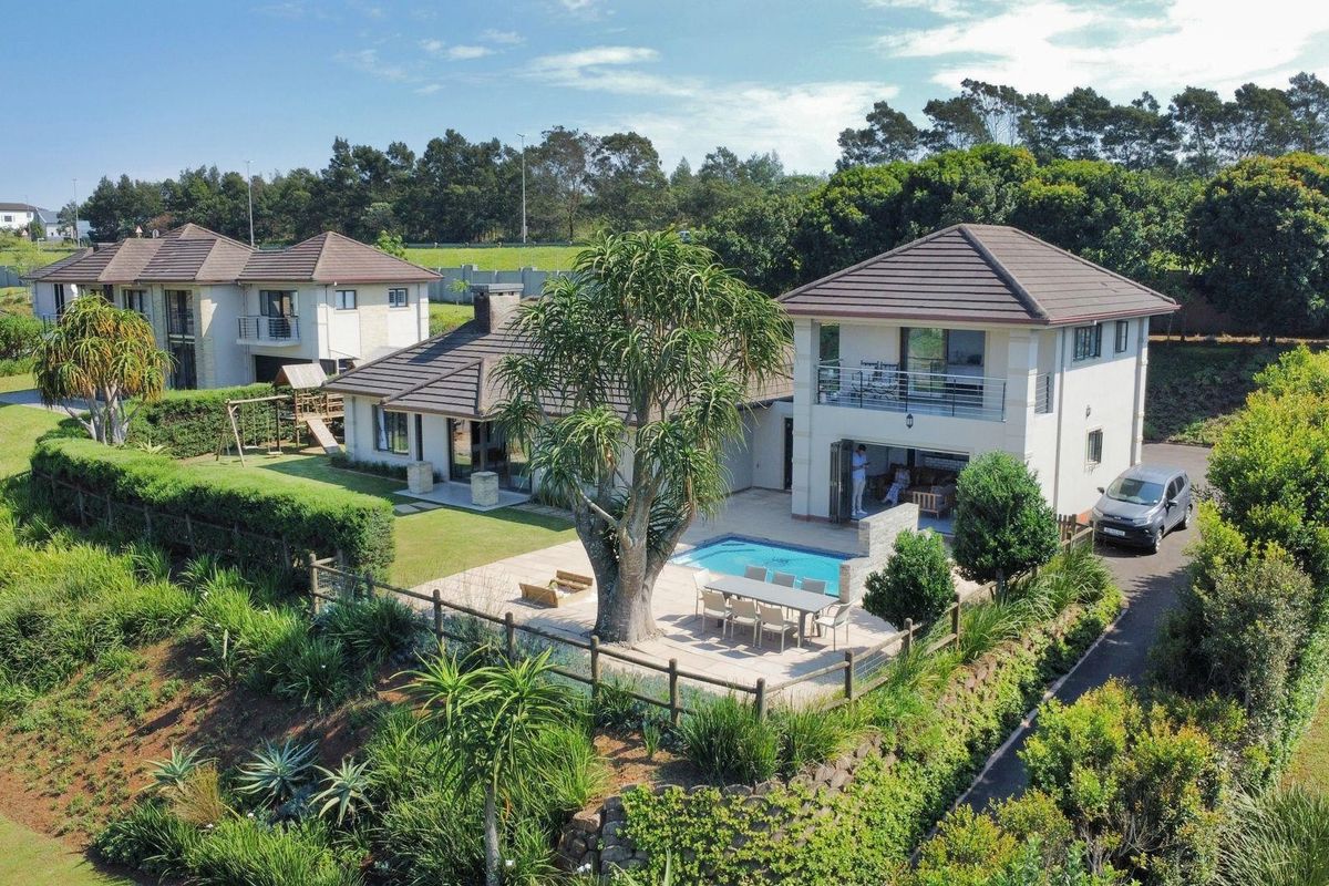 Aerial view of a residential property featuring a swimming pool, paved patio area, landscaped garden, and a multi-level house with a tiled roof.