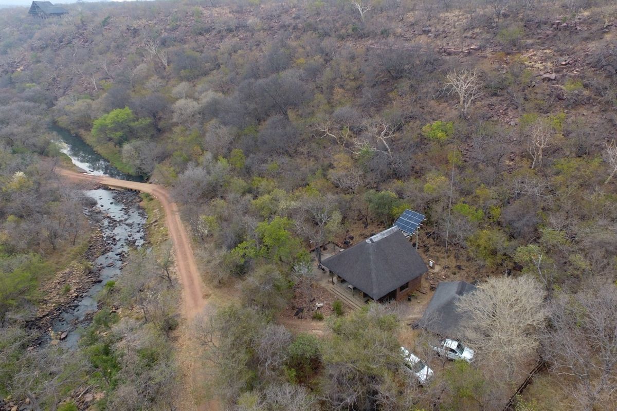 Aerial view of a secluded property featuring a thatched roof house, solar power installation, and natural surroundings with a nearby river and dirt road.