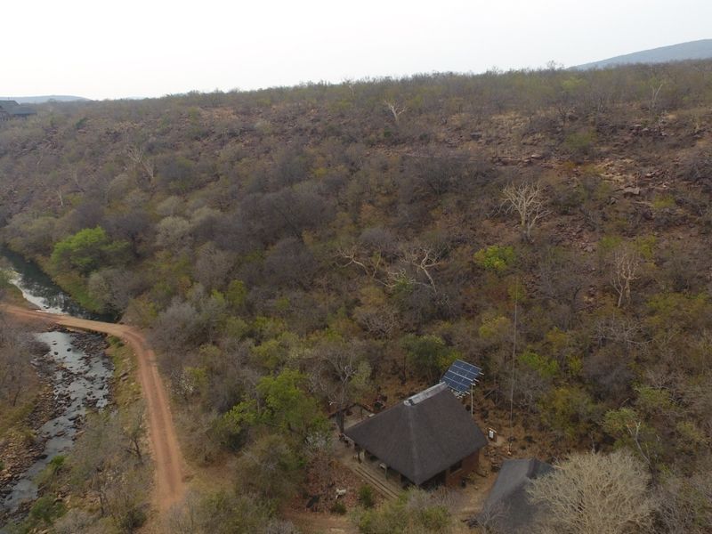 Aerial view of a remote property featuring a thatched-roof structure, solar power installation, and surrounding natural landscape with a nearby stream and dirt road.