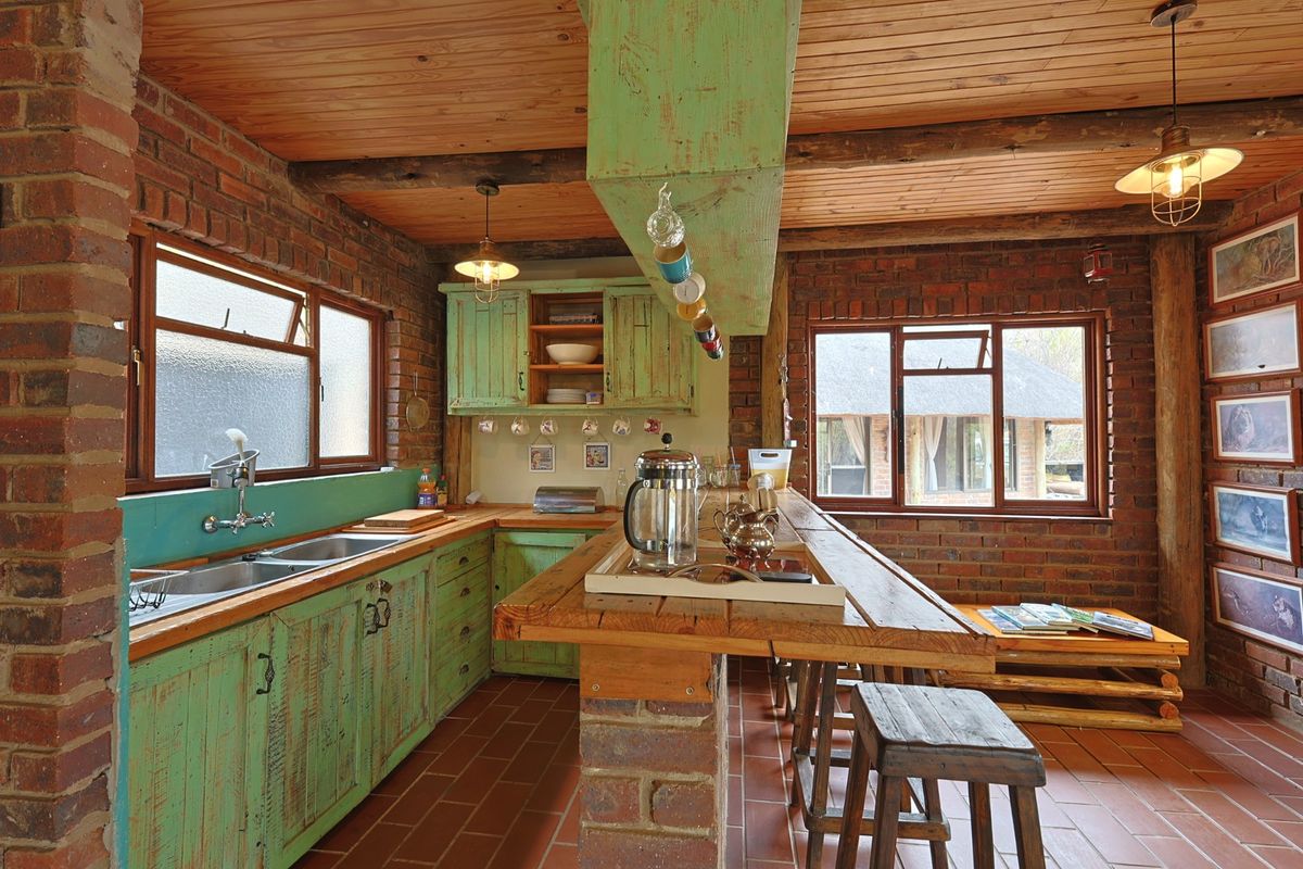 Rustic kitchen featuring exposed brick walls, timber ceiling, fitted green cabinetry, double basin sink, and a central brick breakfast bar with timber top.