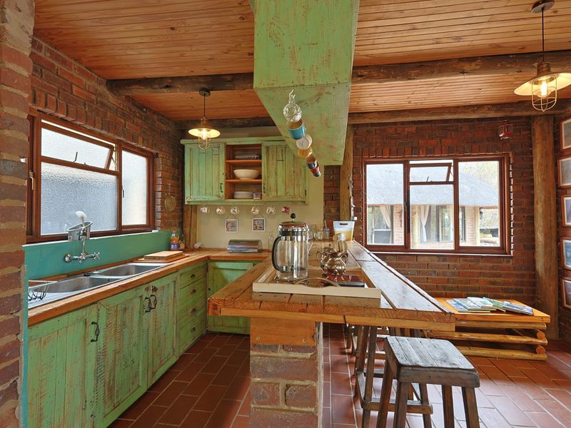 Rustic kitchen featuring exposed brick walls, timber ceiling, fitted green cabinetry, double basin sink, and a central brick breakfast bar with timber top.