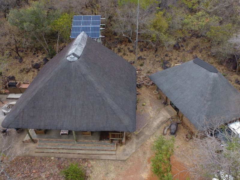 Aerial view of a property featuring two structures with thatched roofs, a solar panel array, and surrounding natural bushveld landscape.