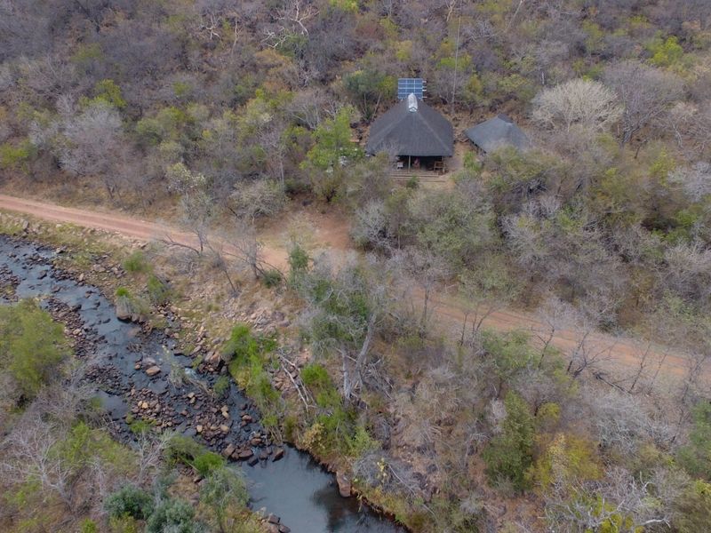 Aerial view of a property featuring a thatched roof dwelling with solar panels, situated in a natural bushveld setting near a river.