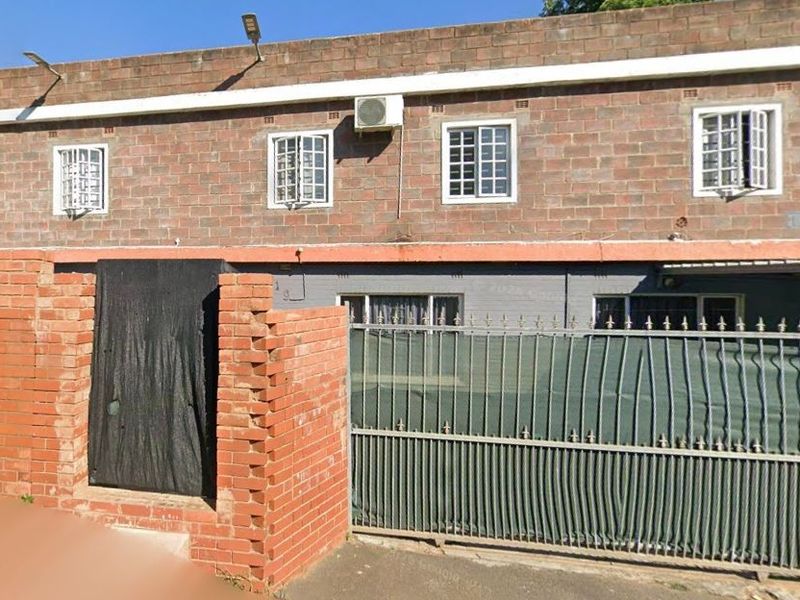 Front elevation of a two-story exposed brick house featuring security bars on windows, a metal sliding gate, and a brick boundary wall.