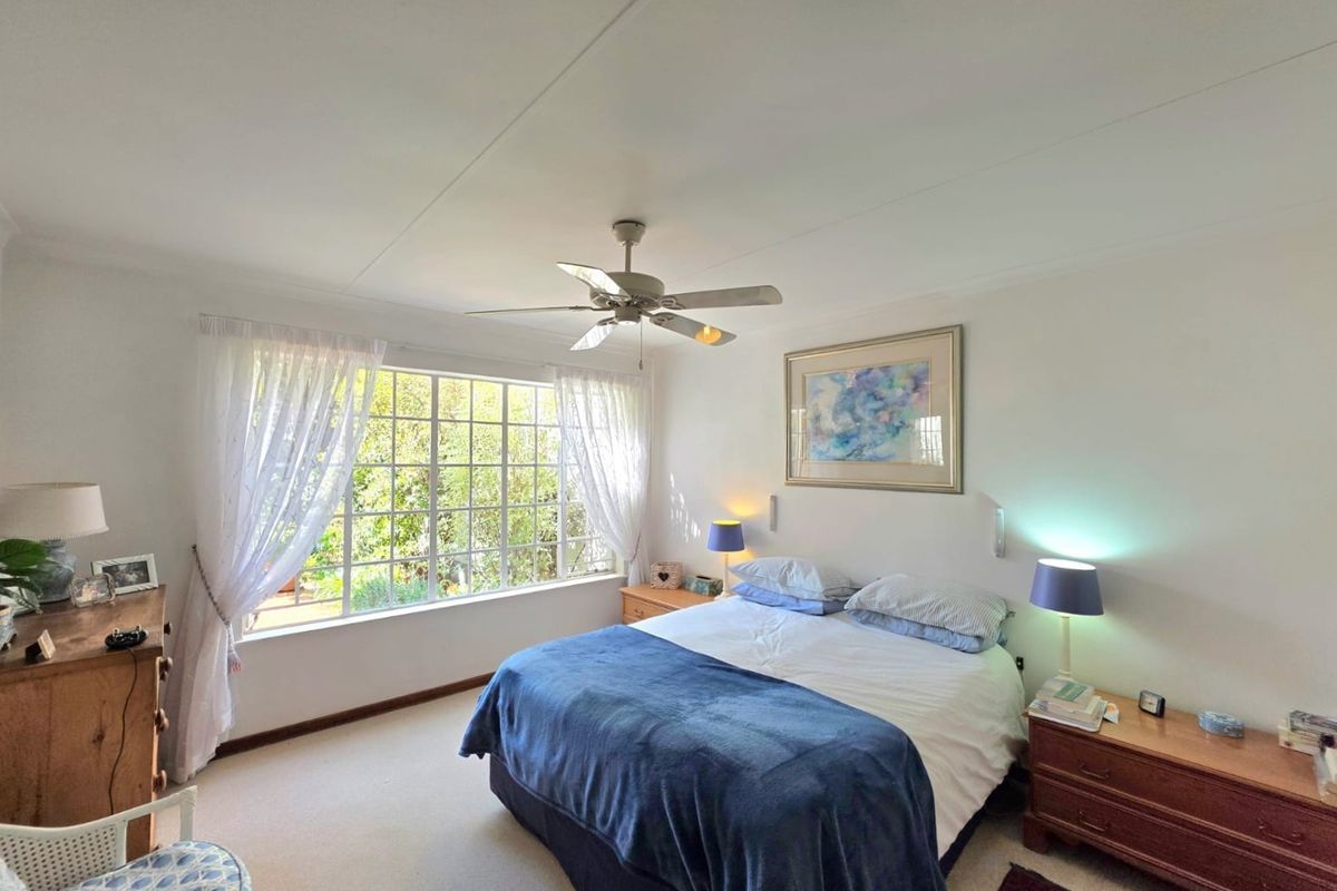 A well-lit bedroom featuring a ceiling fan, large window with natural light, and neutral carpeted flooring.