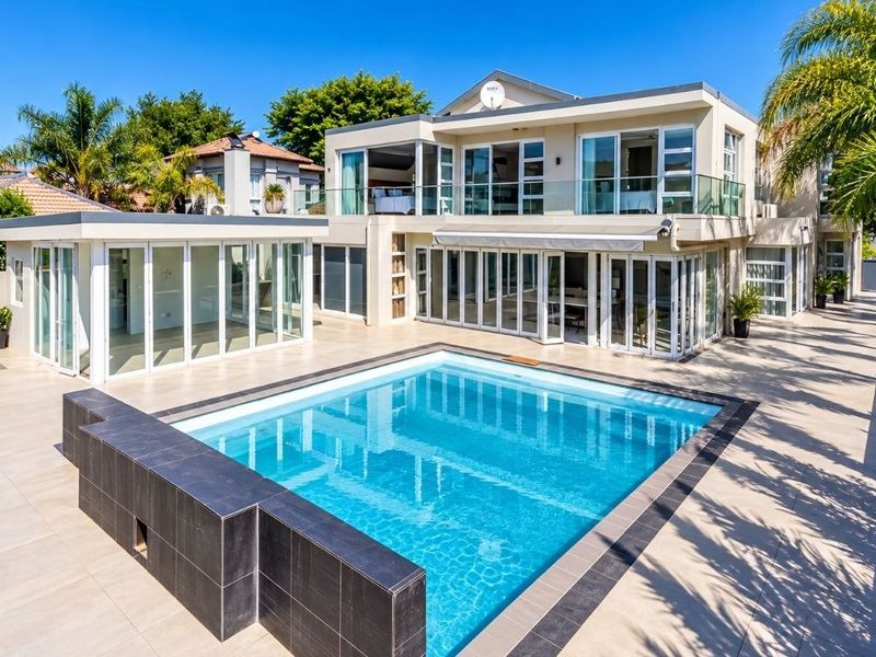 Rear view of a modern double-story house featuring a swimming pool, expansive tiled patio, and large glass sliding doors leading to the interior living spaces.