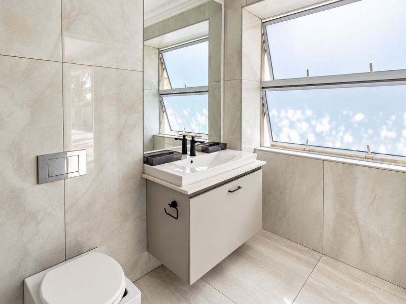 Modern bathroom featuring a wall-mounted vanity, ceramic basin, wall-mounted toilet, and large-format neutral wall tiles with ample natural light from awning windows.