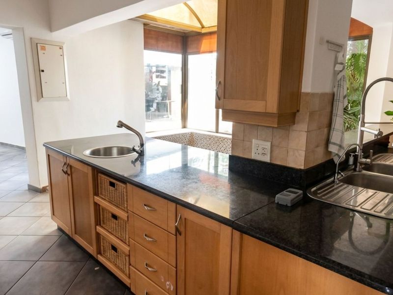 A functional kitchen featuring wooden cabinetry, granite countertops, a double stainless steel sink, and a prep bowl, with tiled flooring and a skylight.