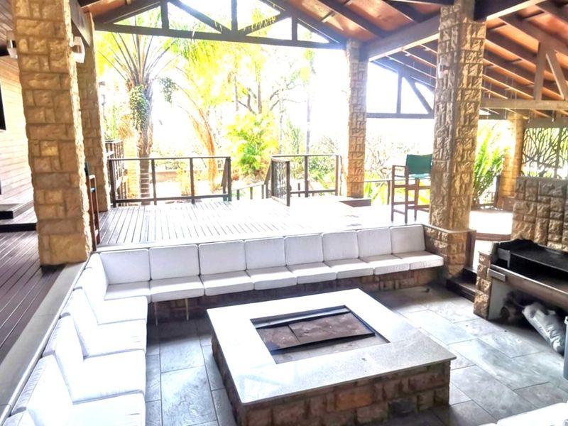 Spacious covered outdoor entertainment area featuring timber decking, a built-in stone braai, and a central stone fire pit with integrated seating.