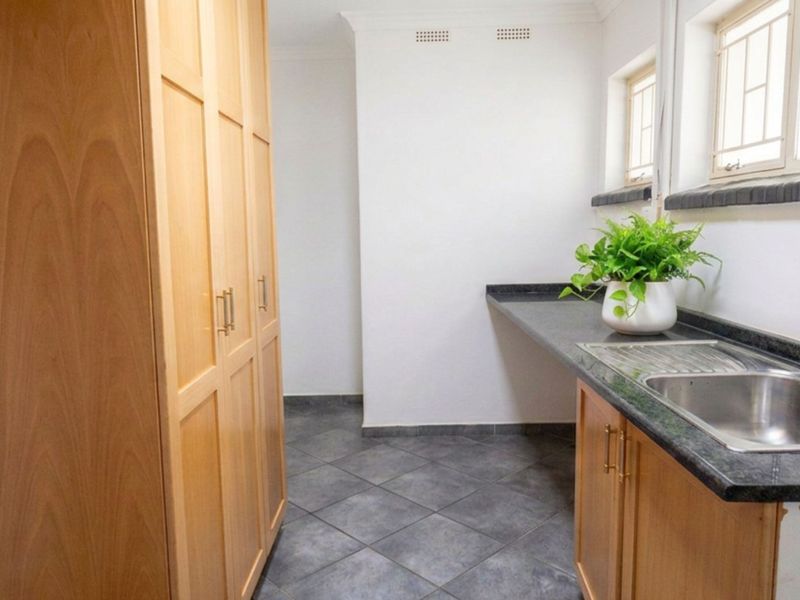 A clean and functional scullery featuring light wood cabinetry, dark stone countertops, a stainless steel sink, and tiled flooring.