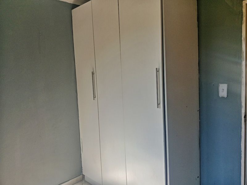 A bedroom interior featuring a built-in white wardrobe against a grey wall, showing signs of wear and unfinished maintenance details.