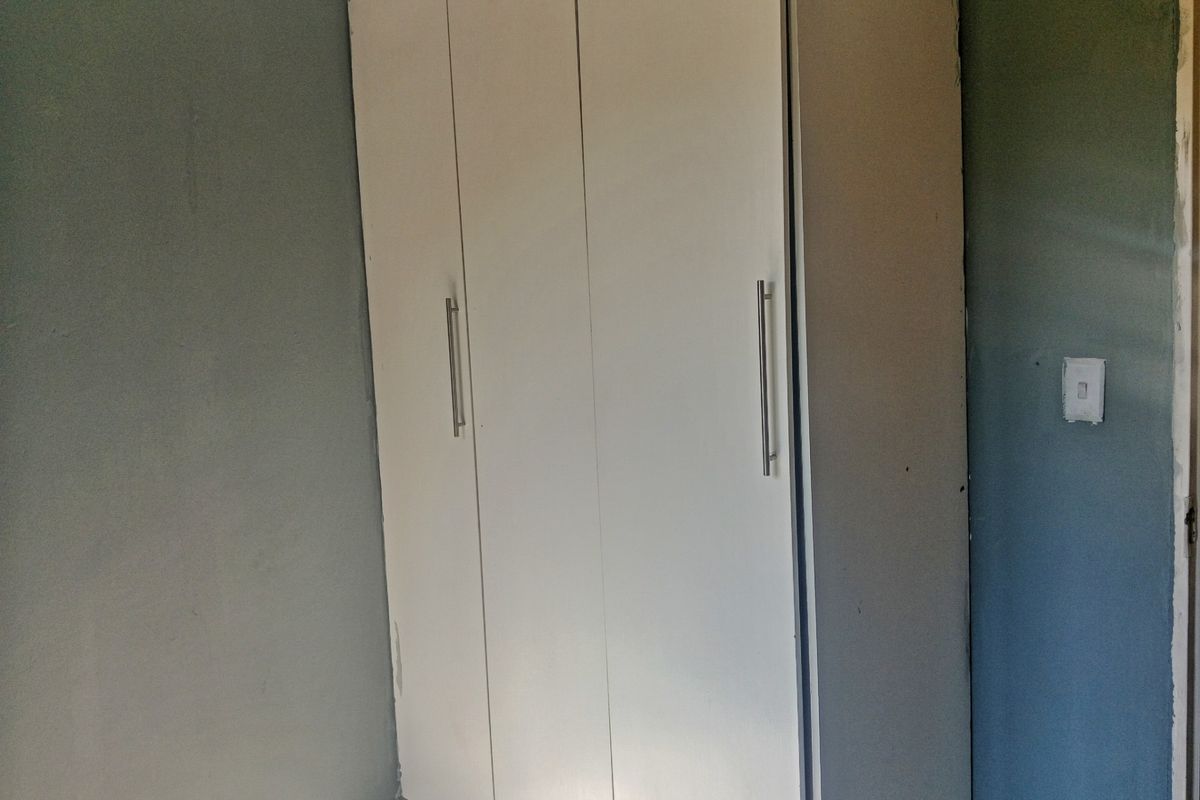 A bedroom interior featuring a built-in white wardrobe against a grey wall, showing signs of wear and unfinished maintenance details.