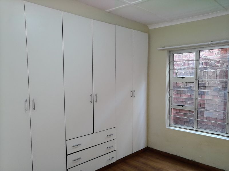 A clean, empty bedroom featuring extensive white built-in cupboards, laminate flooring, and a window fitted with security burglar bars.