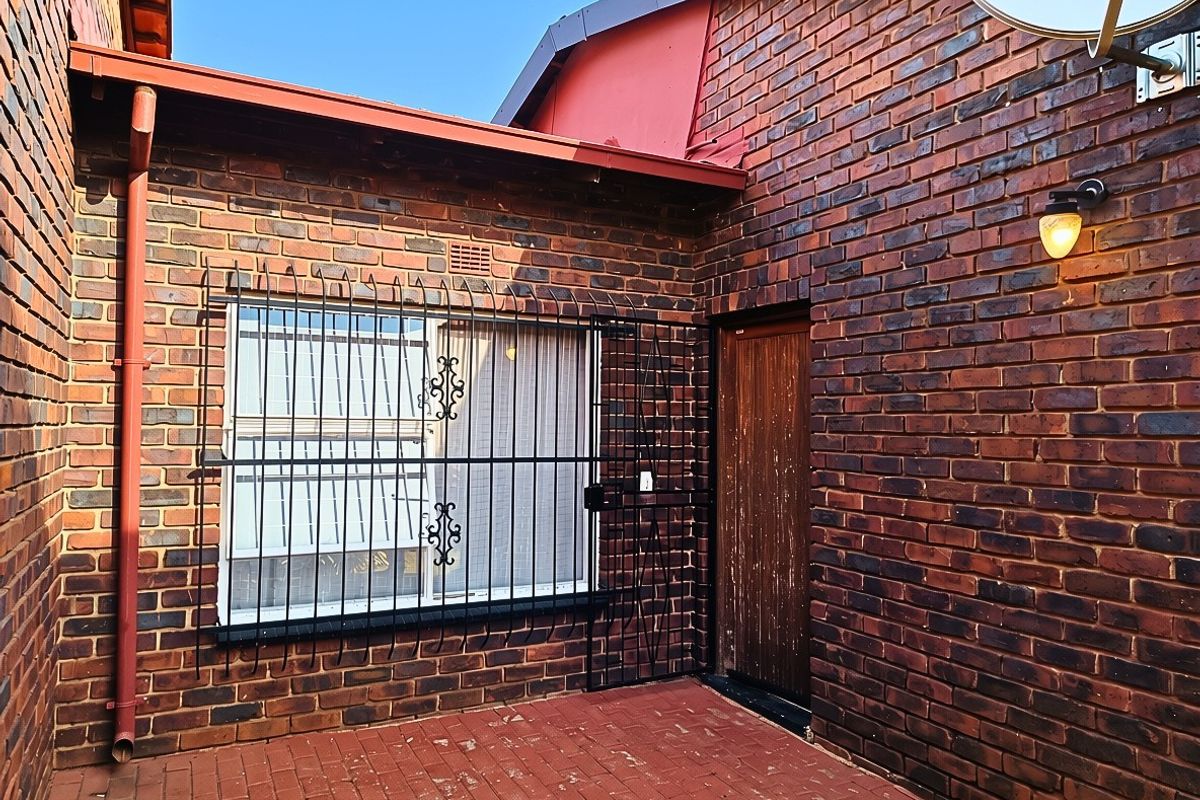 A rear exterior view featuring a face brick facade, paved patio area, secure window bars, and a wooden entrance door.