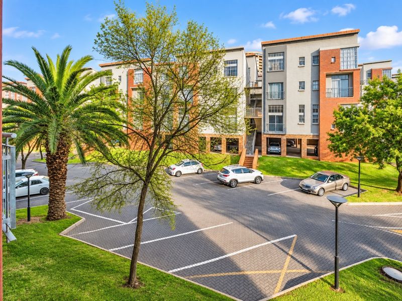 Exterior view of a modern apartment complex featuring a paved parking area, landscaped gardens, and multi-story residential units with balconies.
