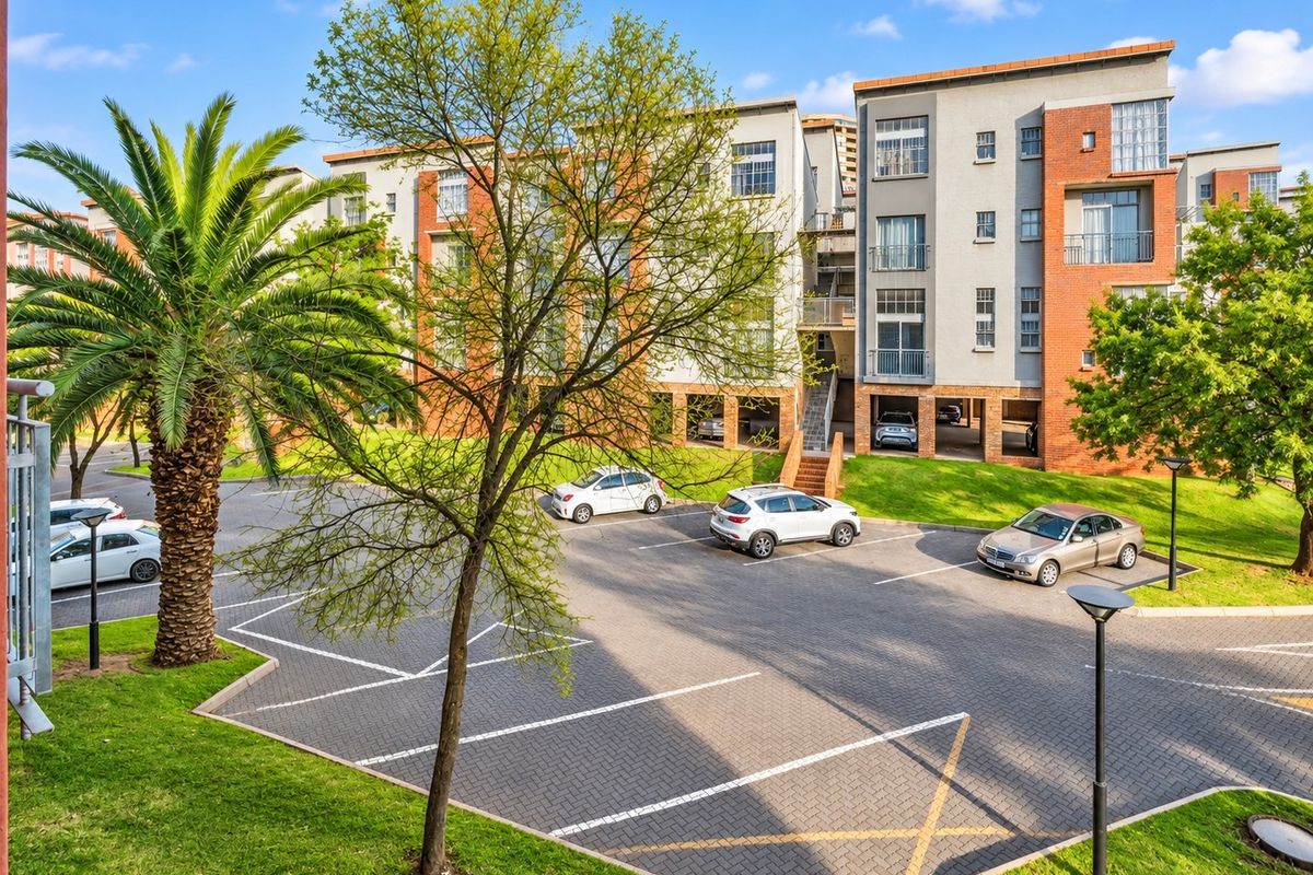 Exterior view of a modern apartment complex featuring a paved parking area, landscaped gardens, and multi-story residential units with balconies.