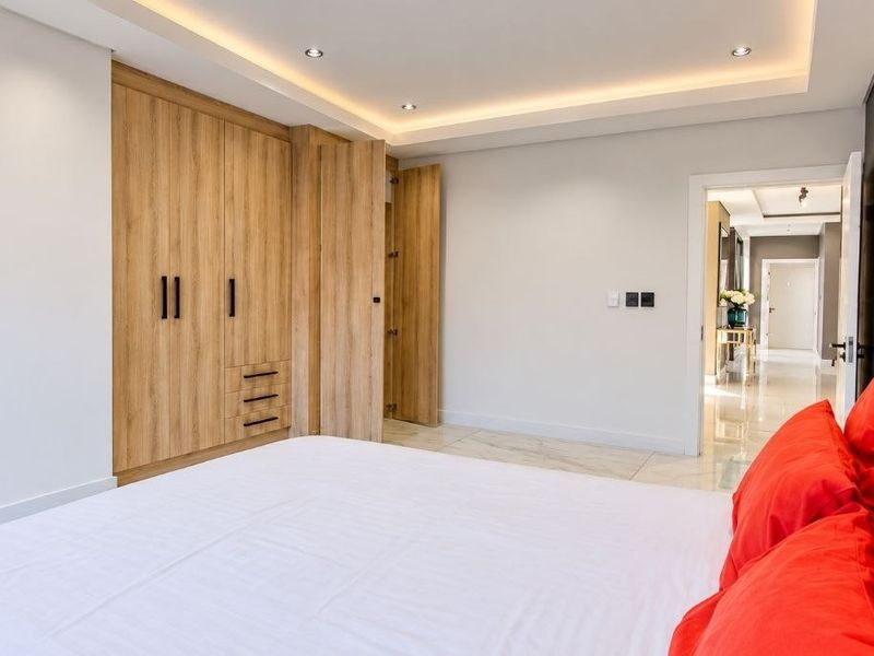 A modern bedroom featuring built-in wooden wardrobes, recessed ceiling lighting with cove details, and polished tiled flooring, leading into a bright hallway.