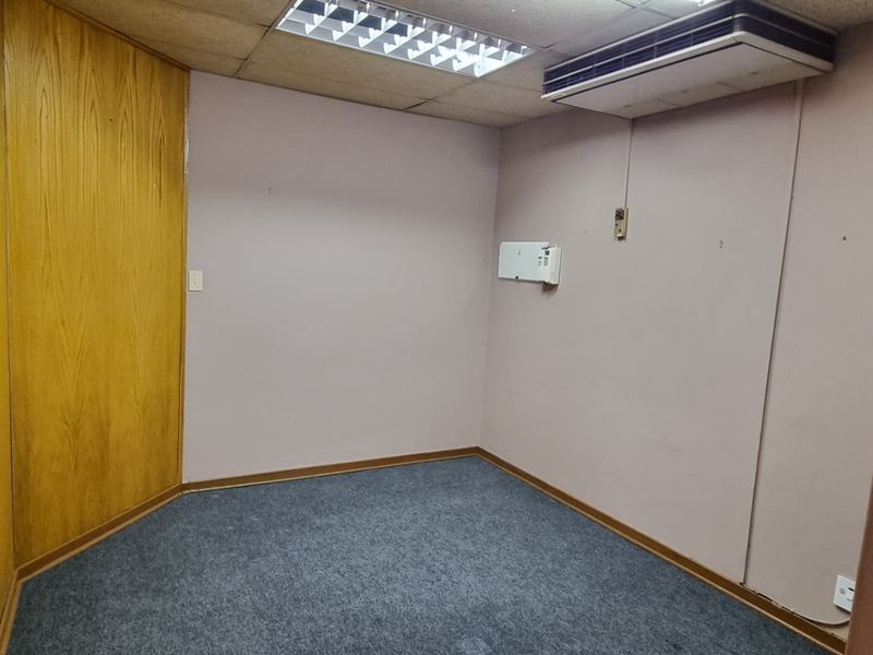 An empty office space featuring wood paneling, carpet flooring, a suspended ceiling with integrated lighting, and a wall-mounted air conditioning unit.