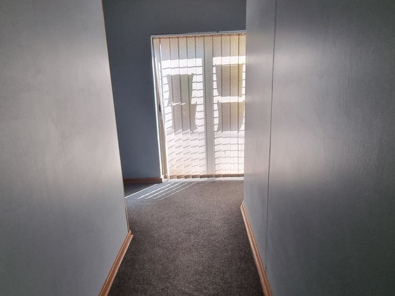 A narrow, empty hallway featuring carpeted flooring and vertical blinds on the window, showing signs of wear on the walls and skirting boards.
