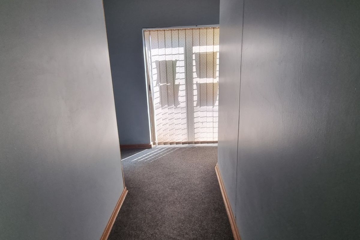 A narrow, empty hallway featuring carpeted flooring and vertical blinds on the window, showing signs of wear on the walls and skirting boards.