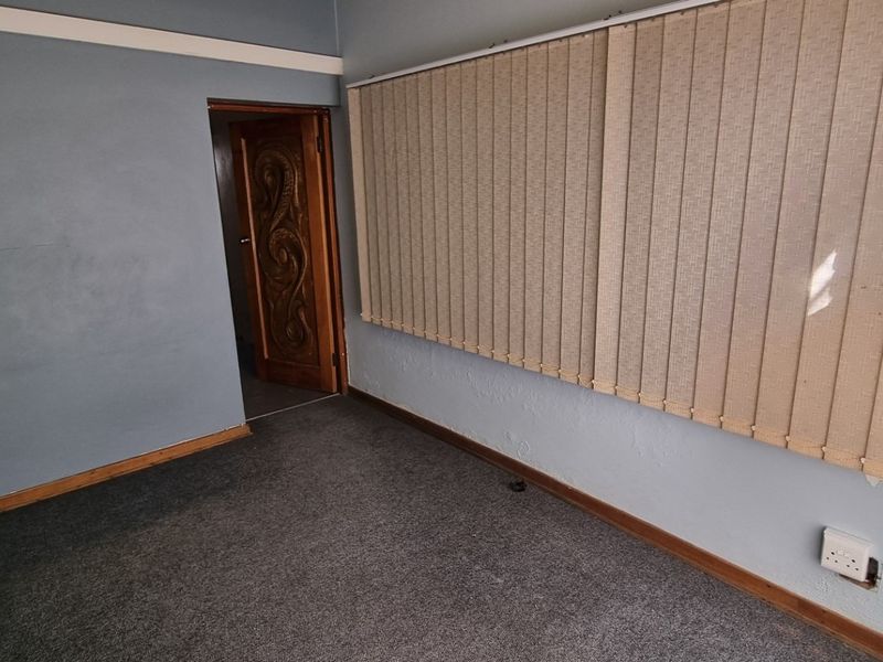 A vacant office space featuring grey carpet flooring, vertical window blinds, and a decorative timber door, requiring minor cosmetic updates.