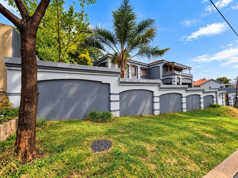 A well-maintained front exterior view of the property featuring a modern grey boundary wall, landscaped lawn, and mature trees.