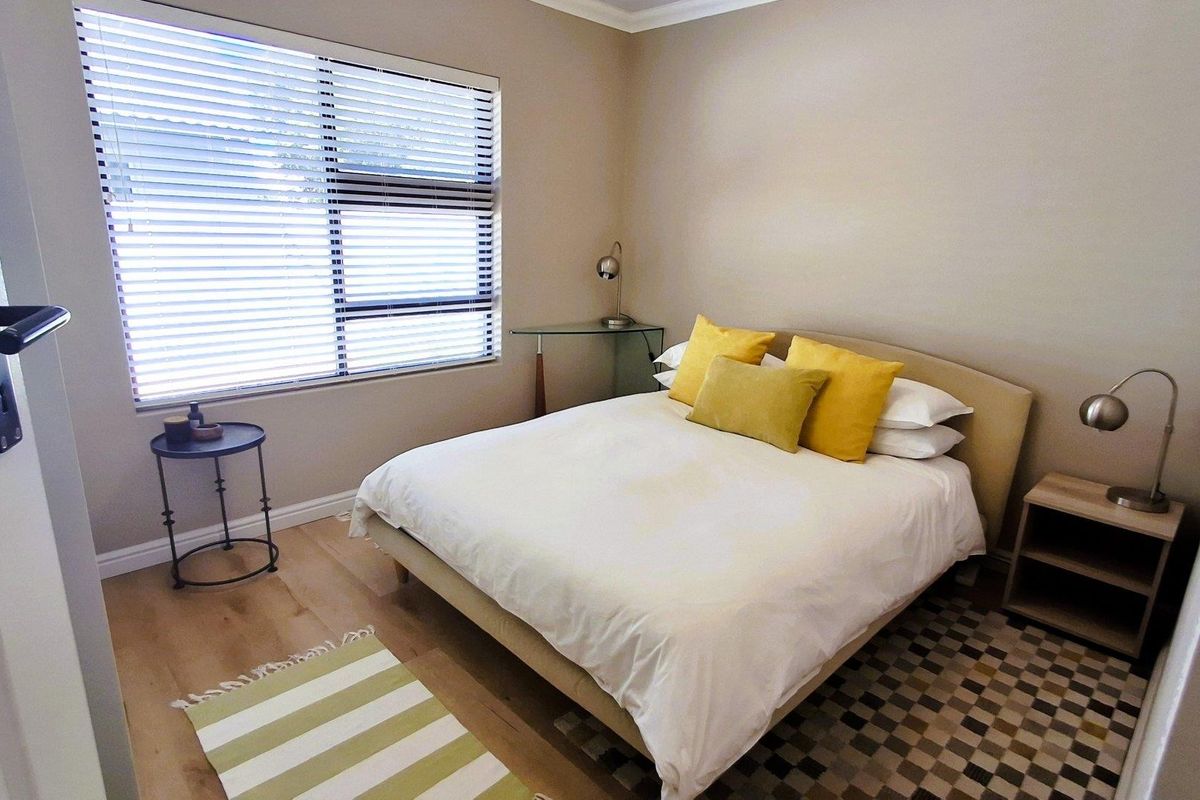 A well-maintained bedroom featuring light-toned laminate flooring, neutral wall finishes, and a large window fitted with venetian blinds.