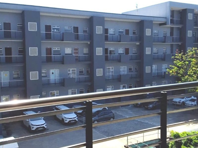 View of a multi-story apartment building facade featuring metal balcony railings and an adjacent paved parking area.