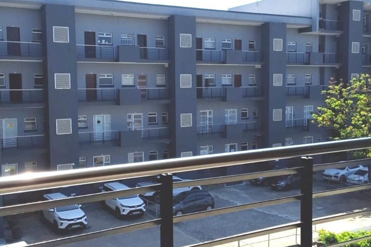 View of a multi-story apartment building facade featuring metal balcony railings and an adjacent paved parking area.