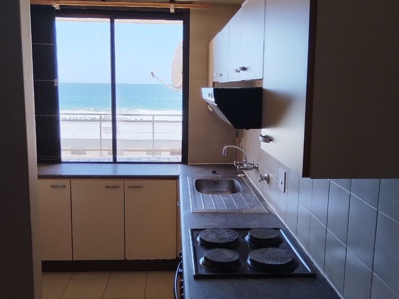 A compact, dated kitchen featuring built-in cabinetry, an electric hob, and a direct view of the ocean through a sliding glass door.