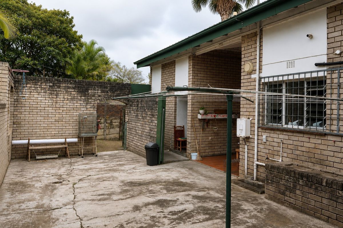 Rear exterior area featuring brick masonry walls, concrete paving, and a covered patio section, showing signs of age and weathering.