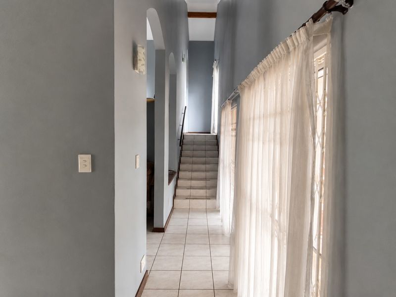 A bright, tiled hallway featuring arched architectural details, a staircase leading to an upper level, and large windows with natural light.