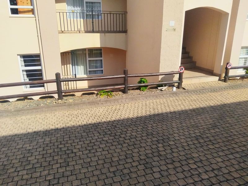 Exterior view of an apartment building entrance featuring a paved driveway, timber fencing, and access to ground floor units and stairwell.