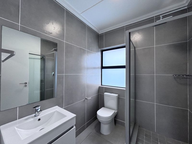 Modern bathroom featuring grey wall and floor tiles, a vanity with basin, a toilet, and a glass-enclosed shower with a rainfall shower head.