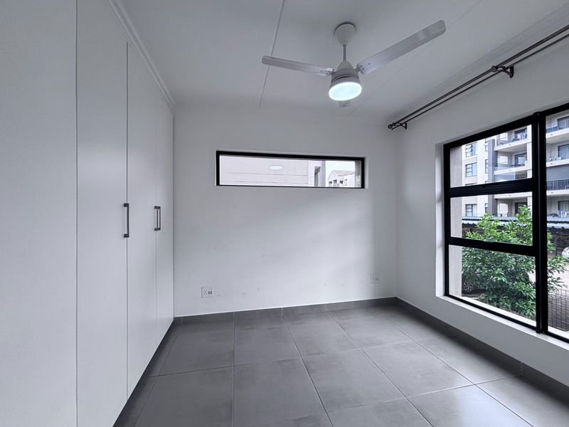 A bright, empty bedroom featuring modern grey floor tiles, built-in cupboards, and a ceiling fan, offering a clean and neutral space.