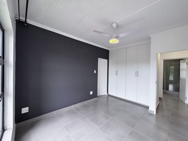 A clean, modern bedroom featuring neutral grey floor tiles, a dark accent wall, built-in storage cupboards, and a ceiling fan.